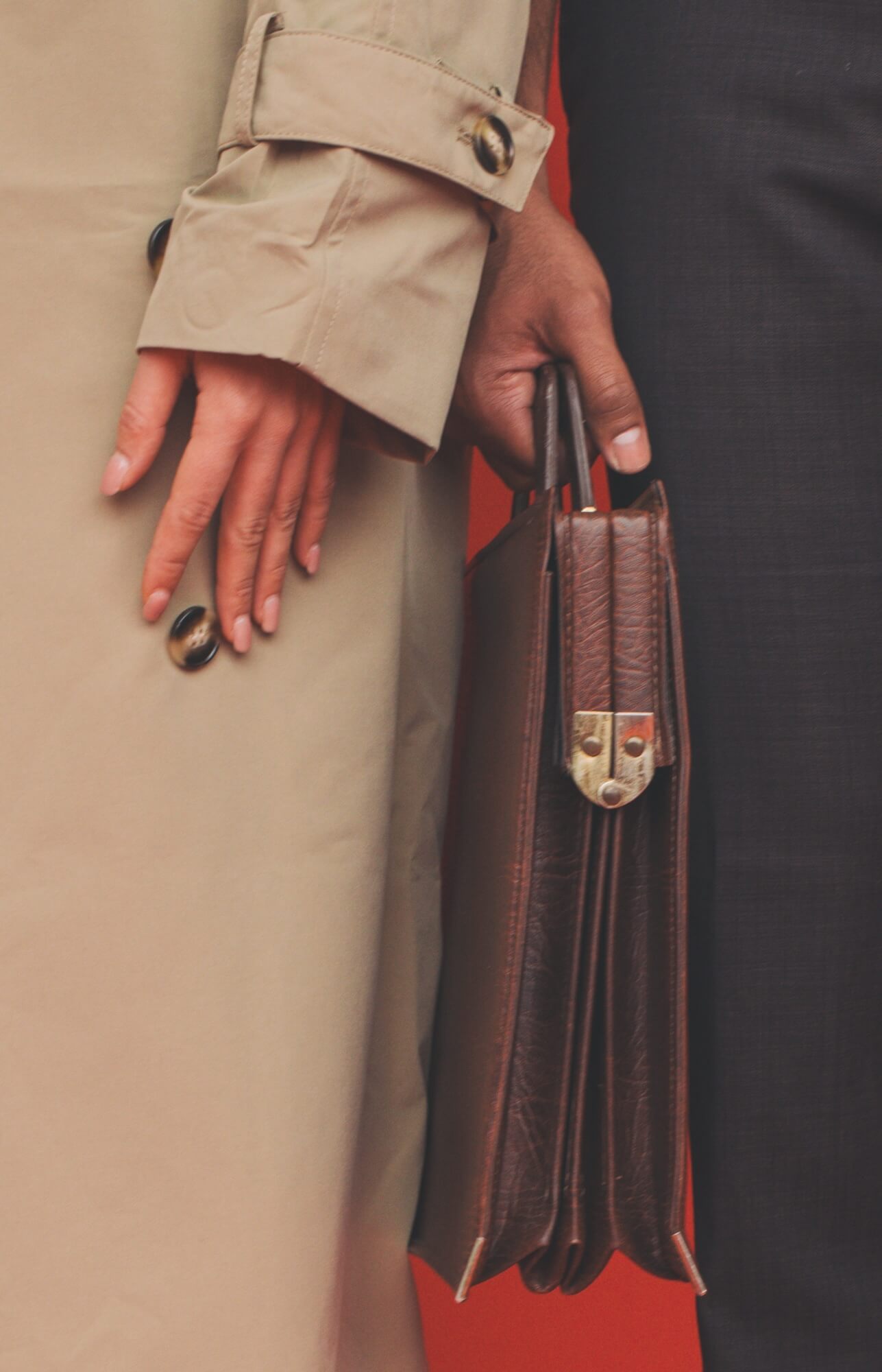 Close-up of two people side by side, one in a beige trench coat and one in a dark suit holding a brown leather briefcase, against a red background.