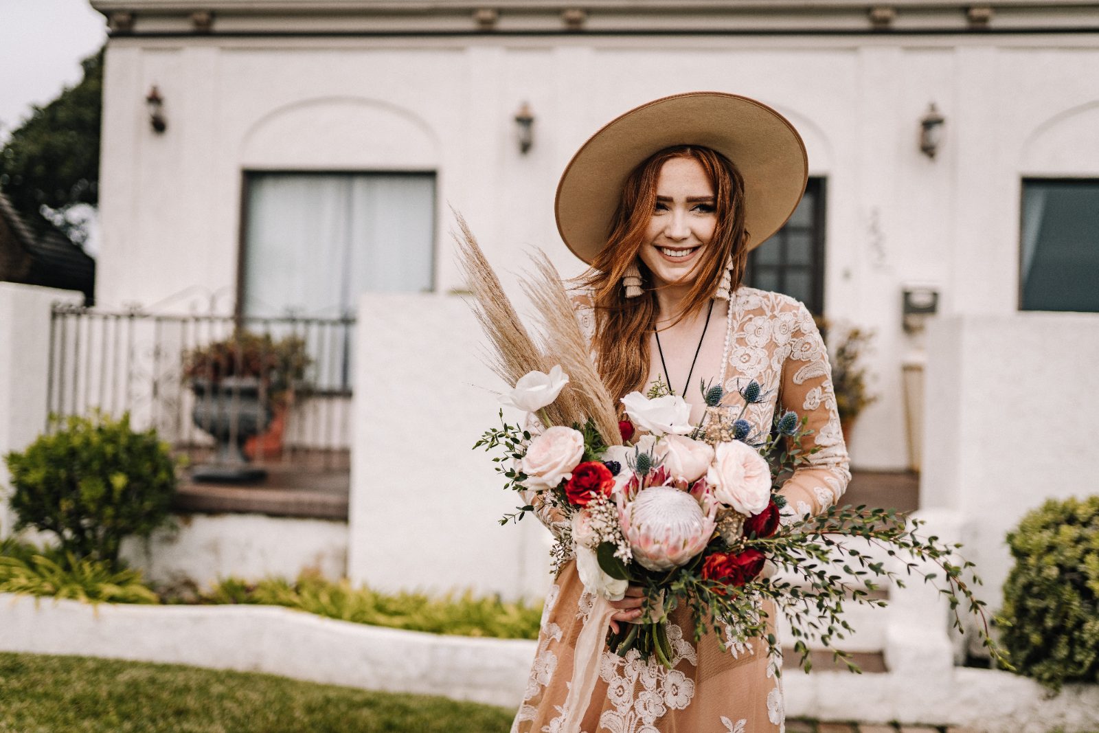 Bride in a wide-brim hat holding a bouquet outside a white venue